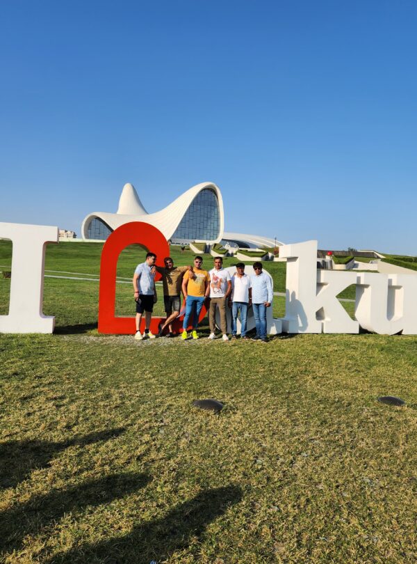 A group of six friends posing in front of the large white "I Love Baku" sign on a grassy hill, with the modern, white Heydar Aliyev Center visible in the background against a clear blue sky.