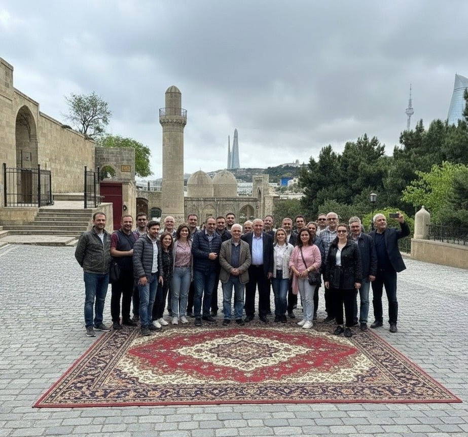 A large group of about 25 people is standing together on a large, red and white traditional Azerbaijani carpet laid over a cobblestone plaza. They are posing in front of an ancient stone mosque and minaret in Baku's Old City. Modern skyscrapers, including the Flame Towers and TV Tower, are visible in the distant background.
