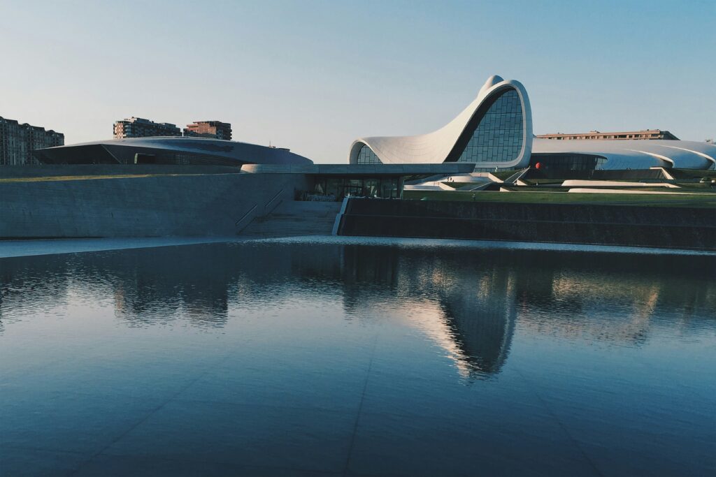 The fluid, wave-like architecture of the Heydar Aliyev Center in Baku, Azerbaijan, is reflected perfectly in a large, dark pool of water in the foreground under a clear sky.