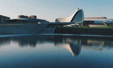 The fluid, wave-like architecture of the Heydar Aliyev Center in Baku, Azerbaijan, is reflected perfectly in a large, dark pool of water in the foreground under a clear sky.Azerbaijan 4 Days & 3 Nights Tour Packages 