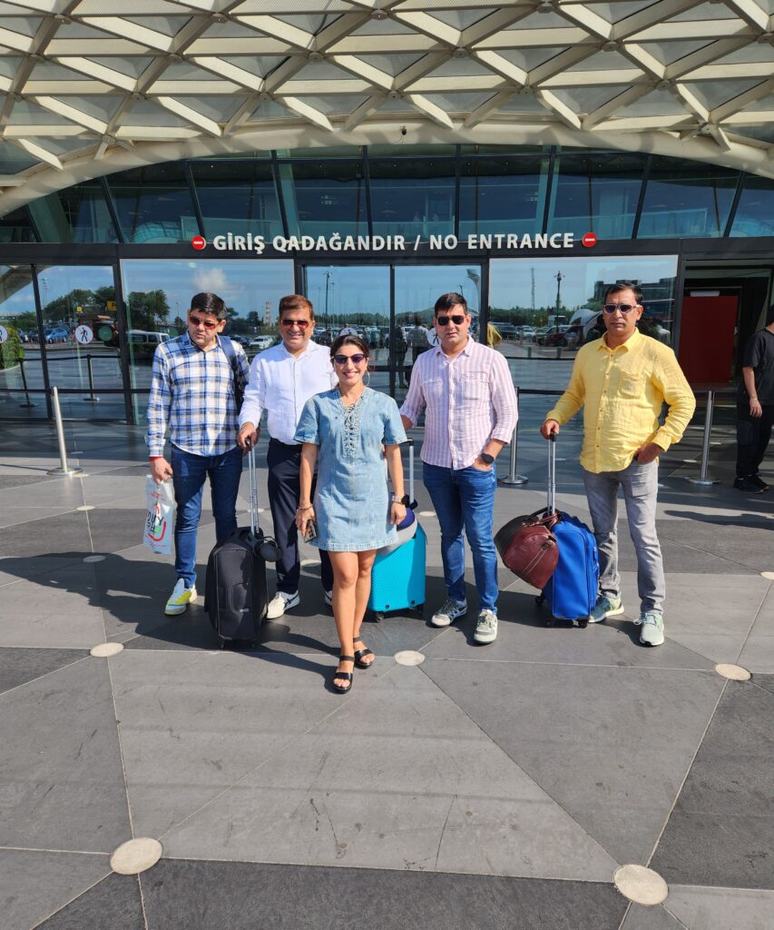 "Five travelers, four men and one woman, standing with luggage outside a modern glass and steel airport terminal with a bilingual sign reading 'GİRİŞ QADAĞANDIR' and 'NO ENTRANCE' — promoting Azerbaijan Tour Packages."