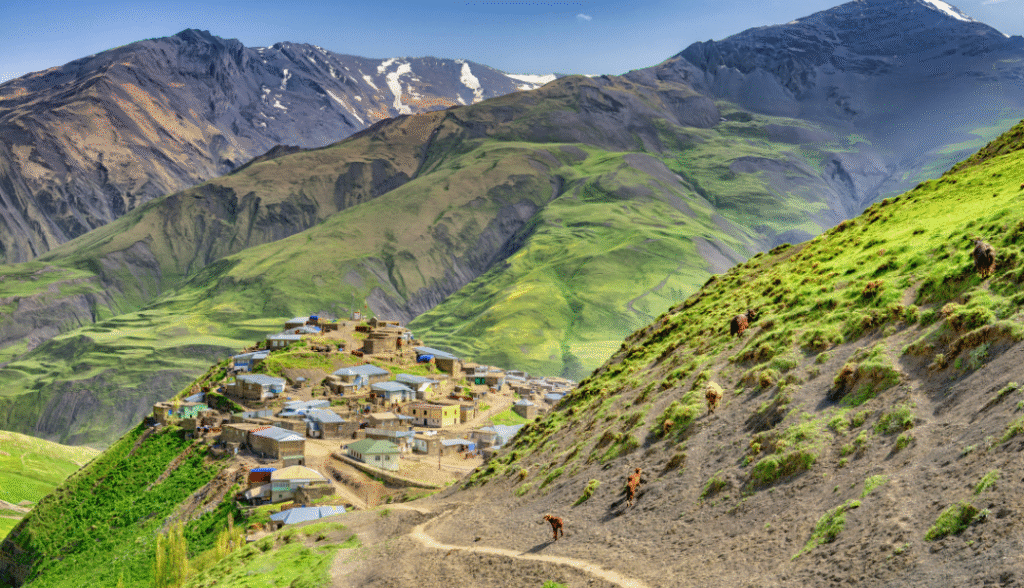 An elevated view of a small, remote mountain village nestled on a steep, green hillside in the foreground. The village is composed of traditional, clustered stone and tin-roofed houses. A winding dirt path leads down the hillside, where several horses or livestock are grazing. Towering, rocky peaks of the Greater Caucasus Mountains, some with patches of snow, dominate the background.