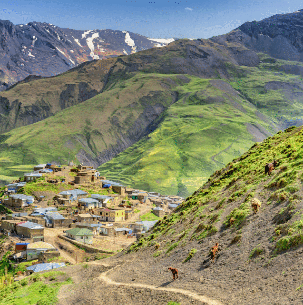 An elevated view of a small, remote mountain village nestled on a steep, green hillside in the foreground. The village is composed of traditional, clustered stone and tin-roofed houses. A winding dirt path leads down the hillside, where several horses or livestock are grazing. Towering, rocky peaks of the Greater Caucasus Mountains, some with patches of snow, dominate the background.