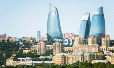 A view of the modern and historic skyline of Baku, Azerbaijan, featuring the three curved, flame-shaped Flame Towers overlooking a cluster of tan, historical buildings and lush green hills.