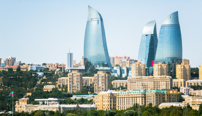 A view of the modern and historic skyline of Baku, Azerbaijan, featuring the three curved, flame-shaped Flame Towers overlooking a cluster of tan, historical buildings and lush green hills.