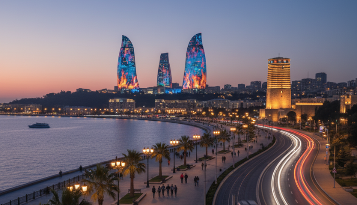 An elevated view of Baku Boulevard (National Park) at dusk, showing a bustling promenade along the Caspian Sea, a road with light trails from vehicles, and the illuminated Flame Towers and other city buildings in the background.