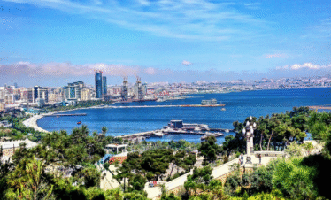 A vibrant, high-angle panoramic view of Baku Bay and the Caspian Sea, showing the modern city skyline, the green parklands of the boulevard, and several ships in the harbor, under a bright blue sky.