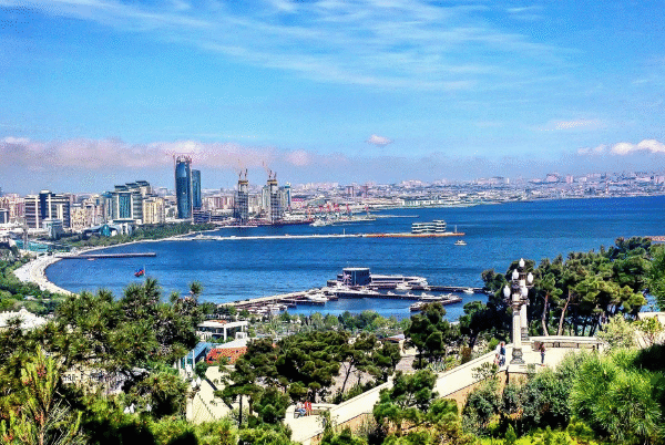 A vibrant, high-angle panoramic view of Baku Bay and the Caspian Sea, showing the modern city skyline, the green parklands of the boulevard, and several ships in the harbor, under a bright blue sky.