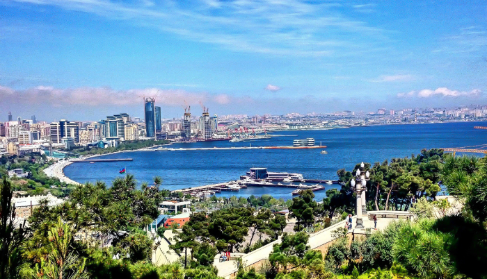 A vibrant, high-angle panoramic view of Baku Bay and the Caspian Sea, showing the modern city skyline, the green parklands of the boulevard, and several ships in the harbor, under a bright blue sky.