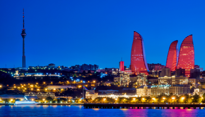 A dramatic night-time panoramic view of the Baku, Azerbaijan skyline, dominated by the three flame-shaped skyscrapers, the Flame Towers, illuminated in vibrant red, and the tall TV Tower on the left.