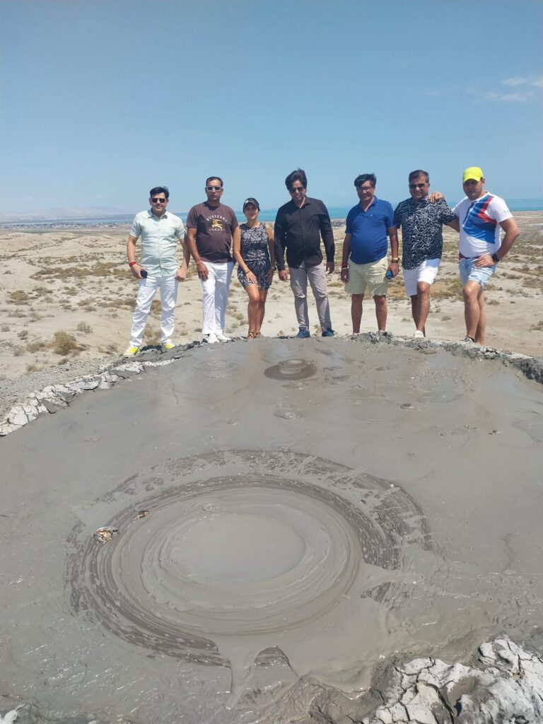 Seven people (six men and one woman) are standing at the rim of a large, circular mud volcano with thick, gray mud in the foreground. They are dressed in casual summer clothing. The background is a dry, rocky, and relatively flat landscape under a bright, clear sky.