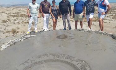 A group of seven adults stands on the rim of a large, circular, gray mud volcano crater, posing for a photo under a bright blue sky in a dry, barren landscape.