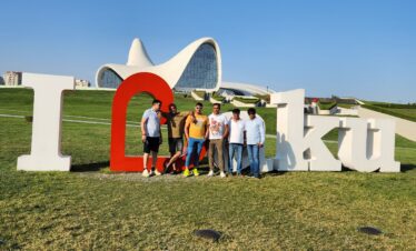 A group of six friends posing in front of the large "I Love Baku" sign on a grassy hill, with the modern, white, wave-like architecture of the Heydar Aliyev Center visible in the background under a clear blue sky.