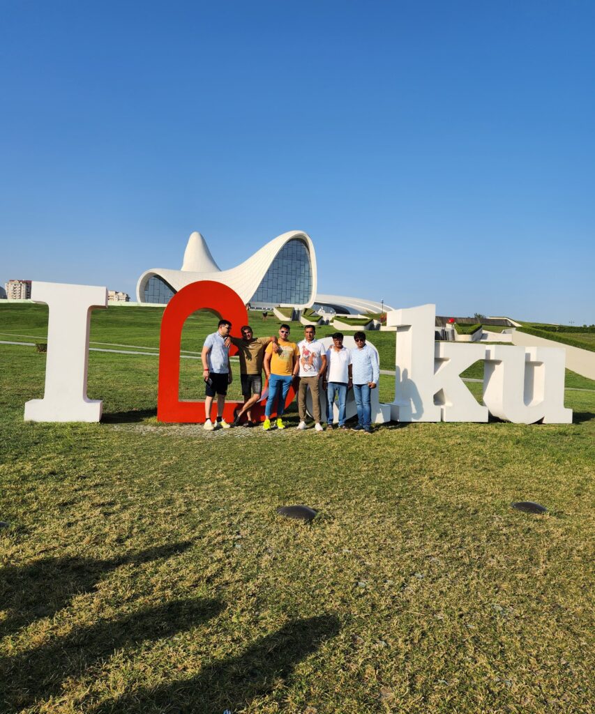 A group of six friends posing in front of the large "I Love Baku" sign on a grassy hill, with the modern, white, wave-like architecture of the Heydar Aliyev Center visible in the background under a clear blue sky.