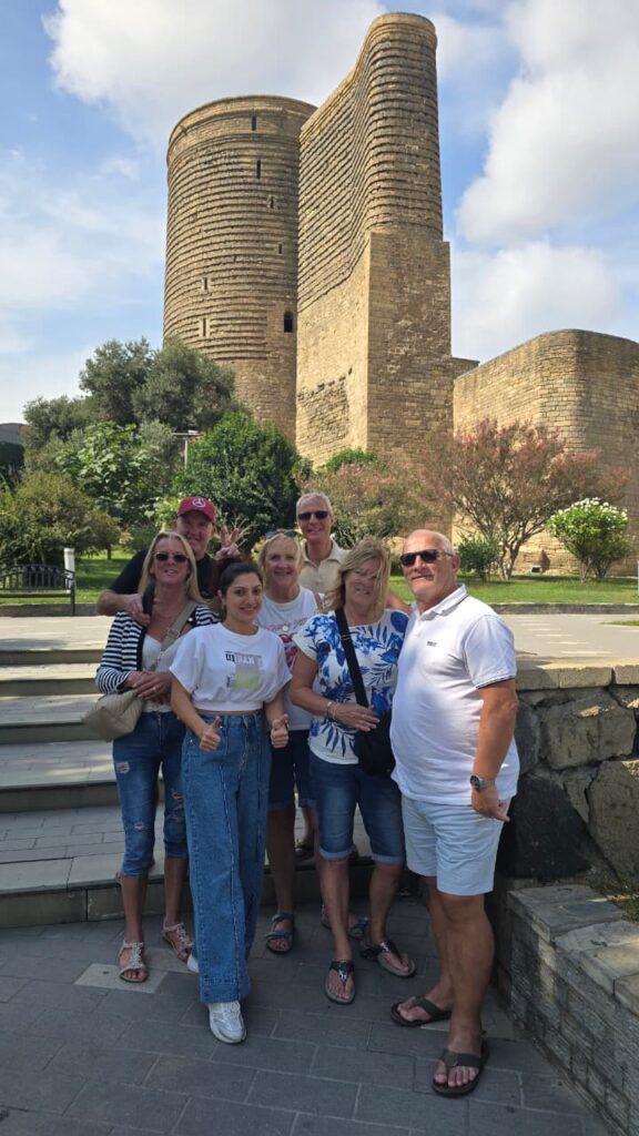 A group of seven tourists smiling and posing together on stone steps in front of the ancient, cylindrical stone structure of the Maiden Tower in Baku, Azerbaijan, under a bright sky.v