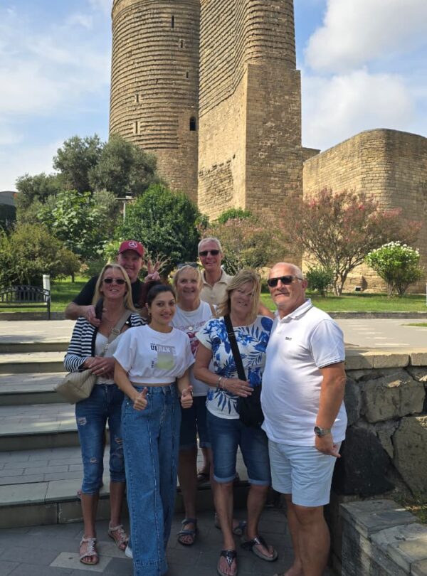 A group of seven tourists smiling and posing together on stone steps in front of the ancient, cylindrical stone structure of the Maiden Tower in Baku, Azerbaijan, under a bright sky.v
