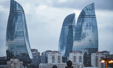 A couple with backpacks, viewed from behind, stands on a bridge looking toward the three futuristic, glass Flame Towers on a cloudy, overcast day, with city buildings below.