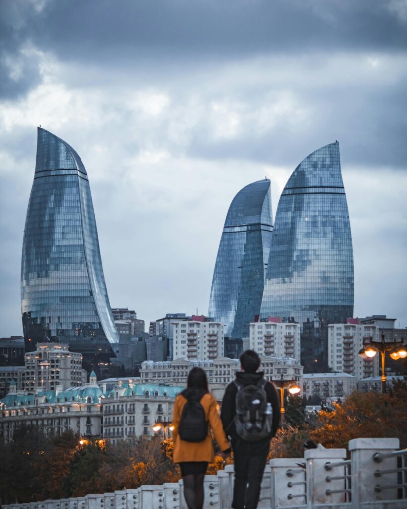 A couple with backpacks, viewed from behind, stands on a bridge looking toward the three futuristic, glass Flame Towers on a cloudy, overcast day, with city buildings below.