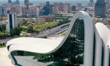 An aerial view of the fluid, wave-like, white architecture of the Heydar Aliyev Center in Baku, with a modern cityscape and the sea visible in the background under a blue sky.