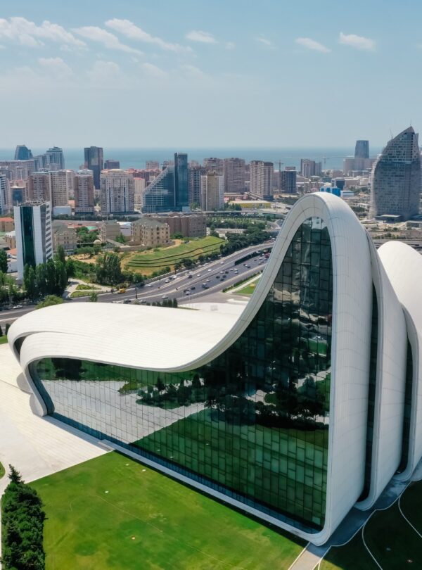 An aerial view of the fluid, wave-like, white architecture of the Heydar Aliyev Center in Baku, with a modern cityscape and the sea visible in the background under a blue sky.