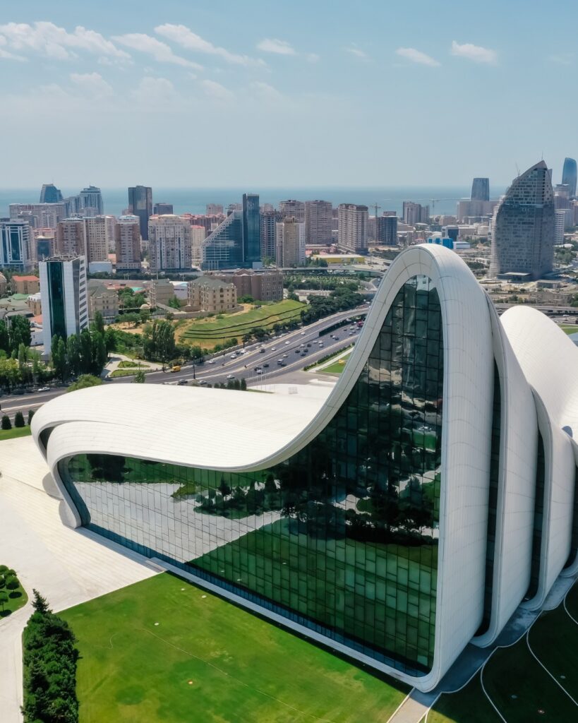 An aerial view of the fluid, wave-like, white architecture of the Heydar Aliyev Center in Baku, with a modern cityscape and the sea visible in the background under a blue sky.