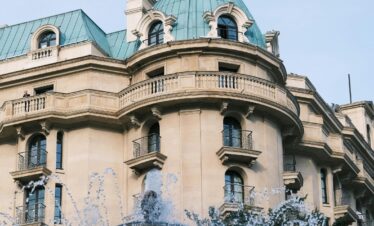 A low-angle shot of a classic European-style building with a beige facade and a distinctive turquoise, domed roof and cupola, partially obscured by the spray of a foreground fountain.