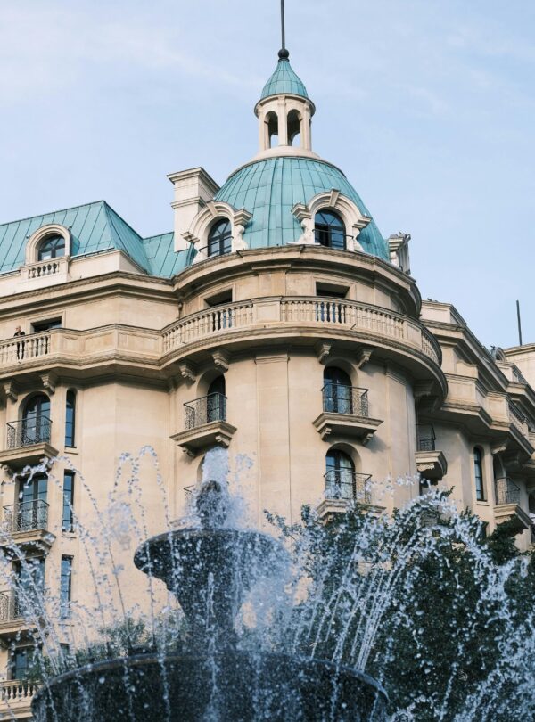 A low-angle shot of a classic European-style building with a beige facade and a distinctive turquoise, domed roof and cupola, partially obscured by the spray of a foreground fountain.