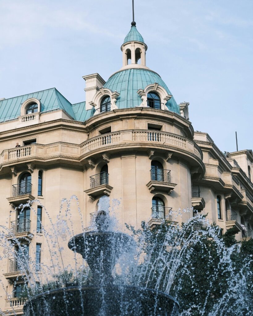 A low-angle shot of a classic European-style building with a beige facade and a distinctive turquoise, domed roof and cupola, partially obscured by the spray of a foreground fountain.