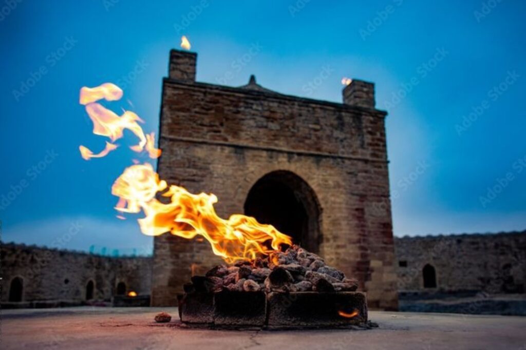 A low-angle shot of a fire burning strongly outside the stone structure of the Ateshgah Fire Temple in Baku, Azerbaijan, under a deep blue sky.