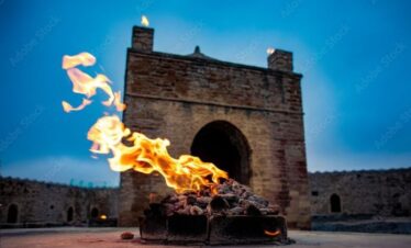 A low-angle shot of a fire burning strongly outside the stone structure of the Ateshgah Fire Temple in Baku, Azerbaijan, under a deep blue sky.
