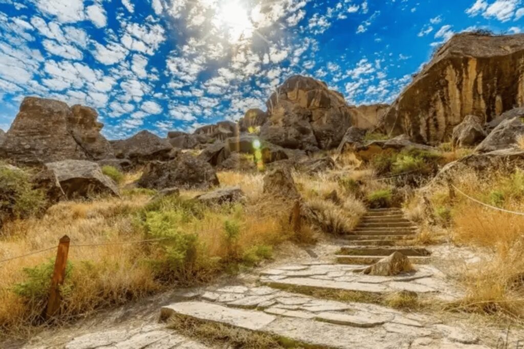 A dramatic landscape featuring rocky, rugged terrain with dry yellow grass and shrubs, traversed by a stone path and steps leading upwards towards large rock formations. The sun is shining brightly under a dynamic blue sky with scattered white clouds.