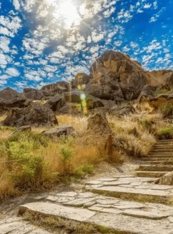 A dramatic landscape featuring rocky, rugged terrain with dry yellow grass and shrubs, traversed by a stone path and steps leading upwards towards large rock formations. The sun is shining brightly under a dynamic blue sky with scattered white clouds.
