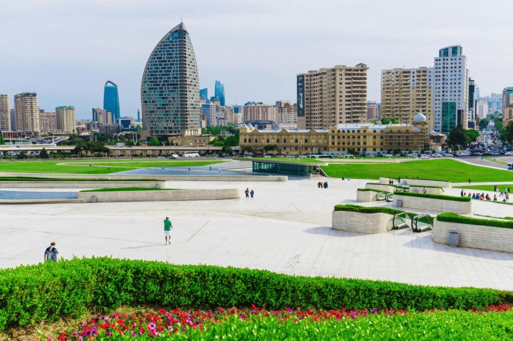 A panoramic view of the modern Baku cityscape featuring a large, bright white plaza with green landscaping and a mix of contemporary high-rise buildings, including the distinctive crescent-shaped tower.