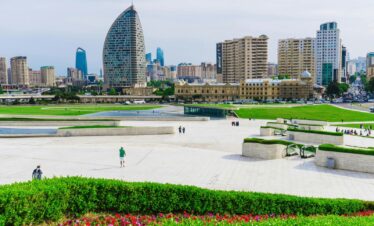 A panoramic view of the modern Baku cityscape featuring a large, bright white plaza with green landscaping and a mix of contemporary high-rise buildings, including the distinctive crescent-shaped tower.Azerbaijan 7 DAYS & 6 NIGHTS TOUR PACKAGES