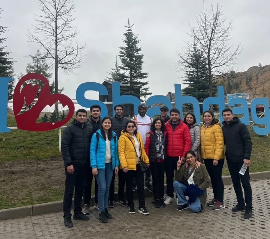 A dramatic natural gas flame burning strongly in the foreground outside the stone central altar structure of the Ateshgah Fire Temple at dusk, contrasting with the dark blue sky.