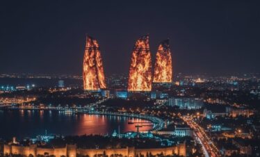 A dramatic night panoramic view of the Baku skyline featuring the tall TV Tower on the left and the three Flame Towers brilliantly illuminated in fiery red against a deep blue sky.