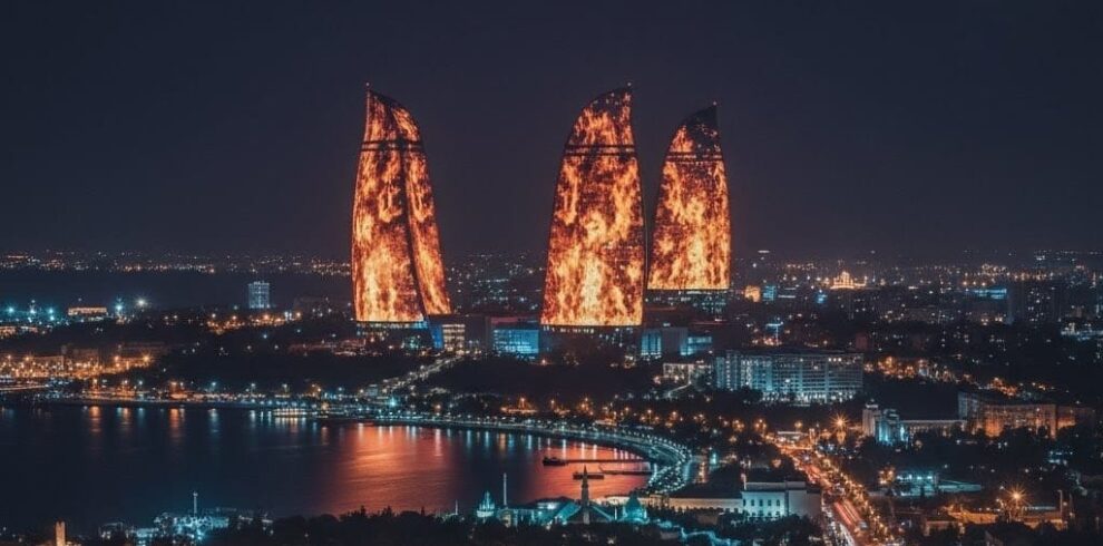 A dramatic night panoramic view of the Baku skyline featuring the tall TV Tower on the left and the three Flame Towers brilliantly illuminated in fiery red against a deep blue sky.