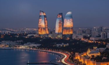 A wide-angle night view of the Baku skyline featuring the tall, illuminated Baku TV Tower on the left and the three Flame Towers brilliantly lit in vibrant red on the right, overlooking the illuminated waterfront.