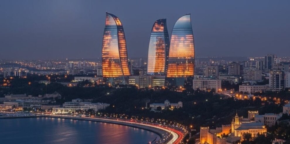 A wide-angle night view of the Baku skyline featuring the tall, illuminated Baku TV Tower on the left and the three Flame Towers brilliantly lit in vibrant red on the right, overlooking the illuminated waterfront.