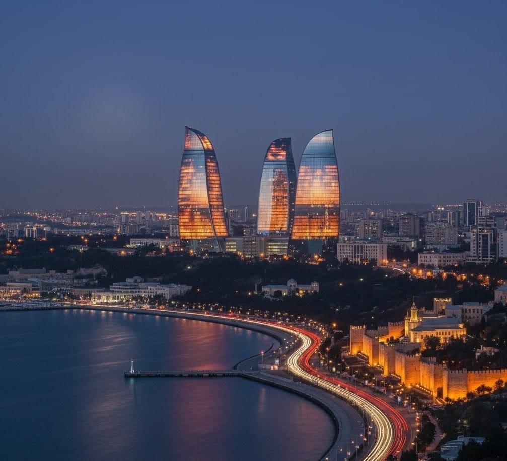 A wide-angle night view of the Baku skyline featuring the tall, illuminated Baku TV Tower on the left and the three Flame Towers brilliantly lit in vibrant red on the right, overlooking the illuminated waterfront.