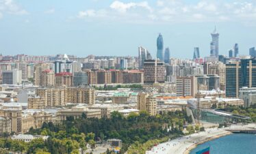 A high-angle panoramic view of a modern coastal city, showing a wide, curved promenade along the turquoise water of the sea. Lush green trees line the foreground and the promenade. The city's skyline features both older, classical buildings and newer, towering skyscrapers, including the distinctive Flame Towers. A large Azerbaijani flag flies prominently near the water.