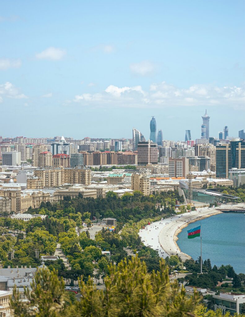 A high-angle panoramic view of a modern coastal city, showing a wide, curved promenade along the turquoise water of the sea. Lush green trees line the foreground and the promenade. The city's skyline features both older, classical buildings and newer, towering skyscrapers, including the distinctive Flame Towers. A large Azerbaijani flag flies prominently near the water.