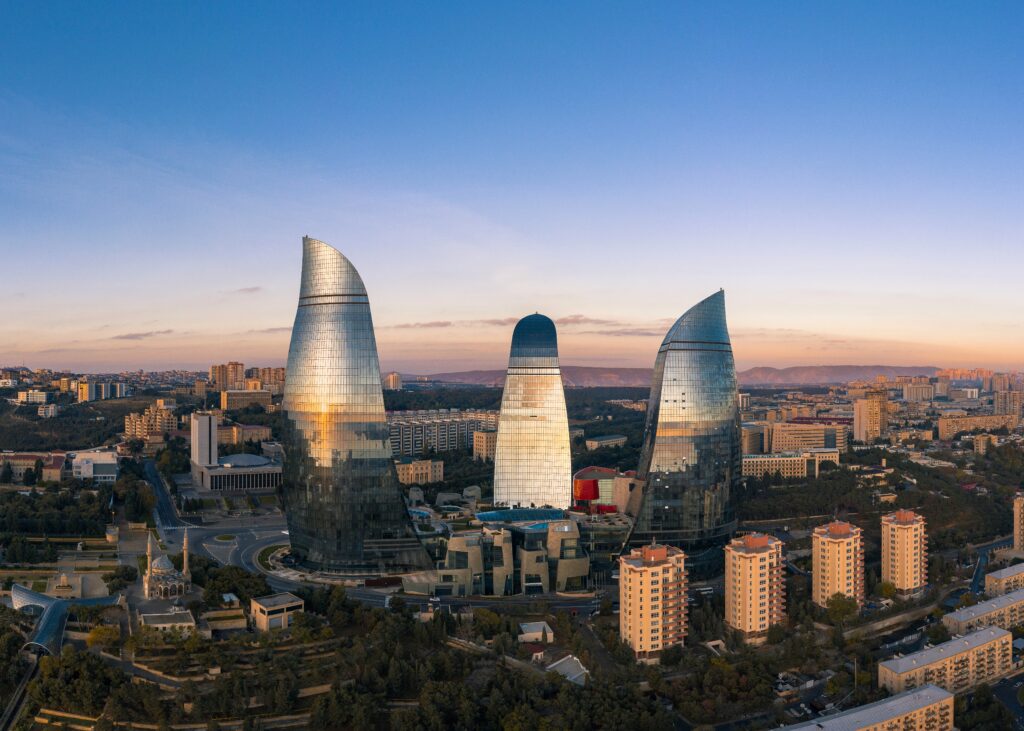 Aerial, wide-angle shot of the three glass, flame-shaped skyscrapers, known as the Flame Towers, dominating the skyline of Baku, Azerbaijan, at dusk. The sky is a gradient of deep blue and orange.