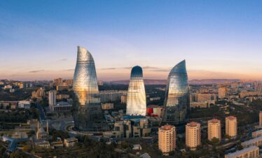Aerial, wide-angle shot of the three glass, flame-shaped skyscrapers, known as the Flame Towers, dominating the skyline of Baku, Azerbaijan, at dusk. The sky is a gradient of deep blue and orange.